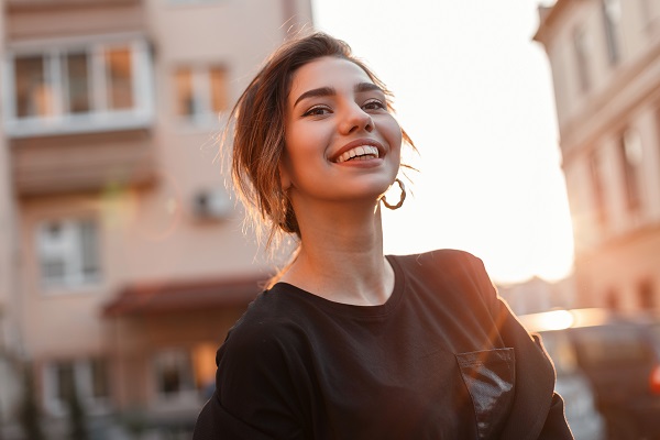 Young happy woman with a beautiful smile in a stylish T-shirt in a coat is standing and enjoying the orange sunset in the city near the houses. Joyful girl model relaxes in the sun. Sunny weekend.