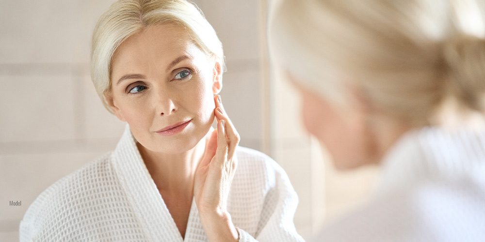 A mature woman in a white robe examines her skin in the mirror.