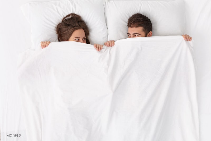 couple in bed with sheets pulled up over their lower faces