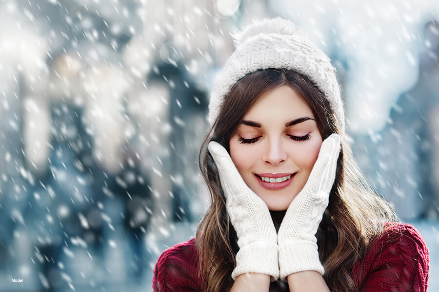 A pretty woman wearing a white hat and gloves holds her face while it's snowing outside.